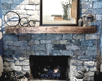 Puede incluir: Una repisa de madera rústica sobre una chimenea de piedra con una pantalla de fuego de metal negro y un inserto de chimenea de gas. La repisa está decorada con tres anillos de metal negro.