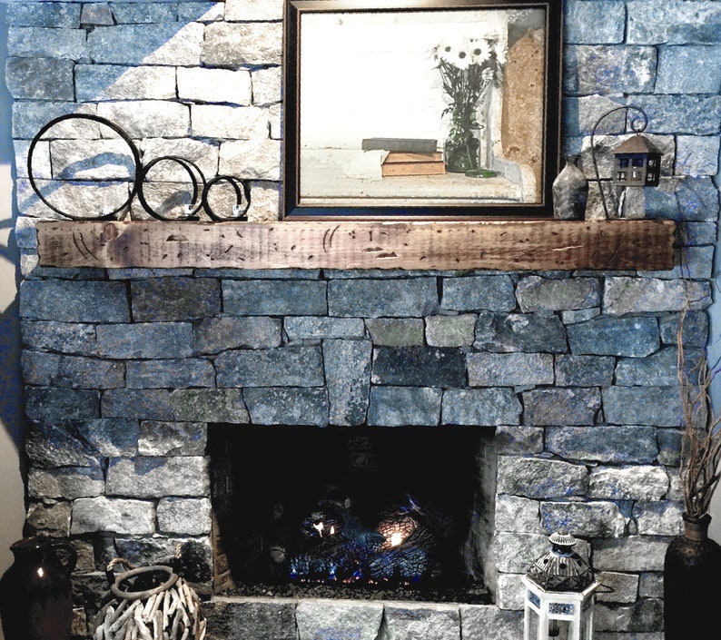 Puede incluir: Una repisa de madera r&uacute;stica sobre una chimenea de piedra con una pantalla de fuego de metal negro y un inserto de chimenea de gas. La repisa est&aacute; decorada con tres anillos de metal negro.