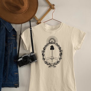 Puede incluir: Una camiseta de color crema con un gr&aacute;fico en blanco y negro de un sol con cara, una mano sosteniendo un palo y un apret&oacute;n de manos dentro de una corona.