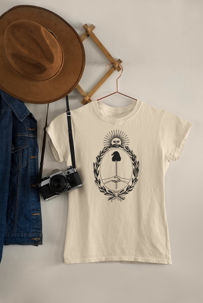 Puede incluir: Una camiseta de color crema con un gr&aacute;fico en blanco y negro de un sol con cara, una mano sosteniendo un palo y un apret&oacute;n de manos dentro de una corona.