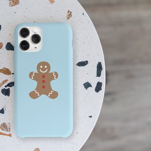 May include: A light blue phone case featuring a gingerbread man design. The gingerbread man is brown with red buttons and a white smile.