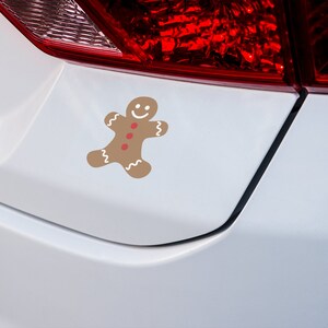 May include: A white car with a gingerbread man sticker on the rear bumper. The gingerbread man is brown with red buttons and a white smile.