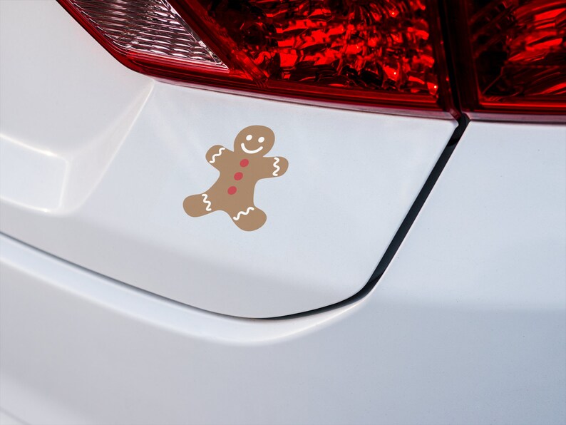 May include: A white car with a gingerbread man sticker on the rear bumper. The gingerbread man is brown with red buttons and a white smile.