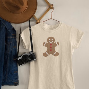 May include: A beige t-shirt featuring a gingerbread man design. The gingerbread man is brown with white icing and red buttons.