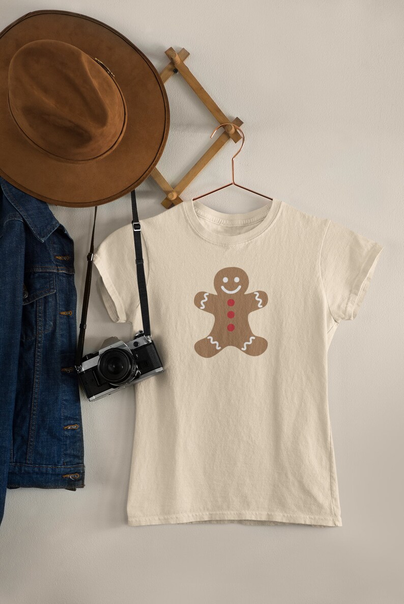 May include: A beige t-shirt featuring a gingerbread man design. The gingerbread man is brown with white icing and red buttons.