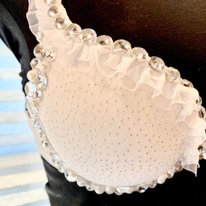 May include: White bra with a ruffled trim and clear rhinestones. The bra is made of a sheer fabric with a dotted pattern.