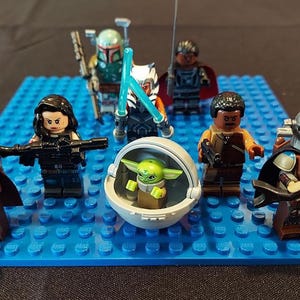 May include: A collection of Lego Star Wars minifigures, including The Mandalorian, Boba Fett, and Ahsoka Tano, arranged on a blue Lego baseplate. The figures are detailed with various weapons and accessories, and a Grogu figure is in a cradle.