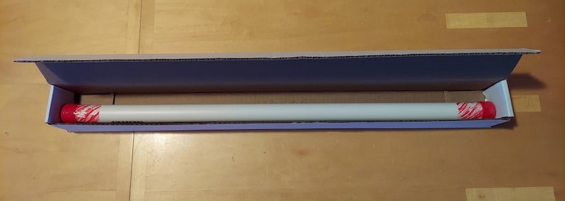 May include: A white cardboard box with the top open, revealing a long, white, cylindrical object with red and white end caps. The object is lying horizontally in the box.