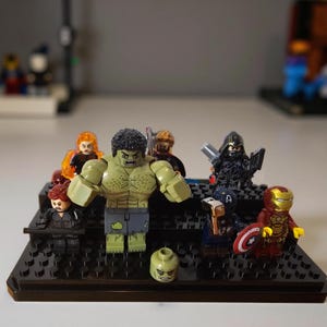 May include: A collection of eight plastic superhero minifigures arranged on a black tiered display stand. The group includes a large green muscular figure, a red and gold armored figure, a blue figure with a shield, and several others in dark outfits.