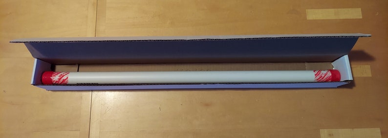 May include: A white cardboard box with the top open, revealing a long, white, cylindrical object with red and white end caps. The object is lying horizontally in the box.