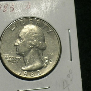 May include: A silver quarter coin from 1983, featuring a profile of George Washington. The coin is in a protective holder with the year 1983 written in red ink at the top. The coin's inscription includes "LIBERTY" and "IN GOD WE TRUST".