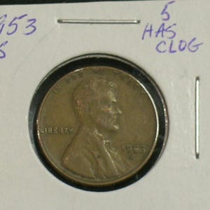 May include: A 1953 Lincoln penny in a white cardboard holder. The coin is bronze-colored and features a profile of Abraham Lincoln. Handwritten text in blue ink above the coin reads "1953" and "5 HAS CLOG".
