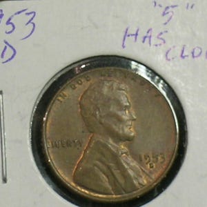 May include: A copper-colored 1953 D Lincoln penny in a protective holder. The obverse of the coin features a profile of Abraham Lincoln. Handwritten text above the coin reads "1953 D" and "5" HAS CLOG".