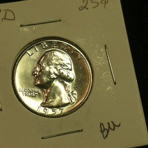 May include: A 1957 D Washington quarter dollar coin encased in a square cardboard holder. The silver-colored coin features the profile of George Washington facing left, with the text LIBERTY, IN GOD WE TRUST, and the year 1957 clearly visible.