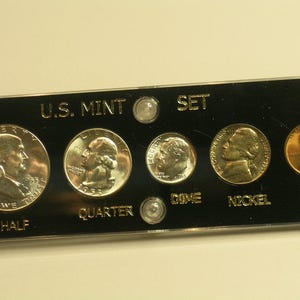 May include: A U.S. Mint set of coins displayed in a black holder. The set includes a half dollar, quarter, dime, nickel, and cent. The coins are silver and gold in color, with the words "U.S. MINT SET" printed on the holder.