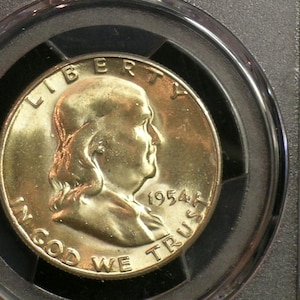May include: A gold-colored coin featuring a profile of a man, with the inscription "LIBERTY" at the top and "IN GOD WE TRUST" at the bottom. The year "1954" is visible. The coin is encased in a clear protective holder.