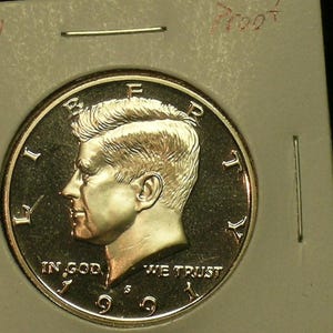 May include: A 1991 S Kennedy half dollar proof coin. The coin features a profile of John F. Kennedy and the inscriptions "LIBERTY", "IN GOD WE TRUST", and "WE TRUST". The coin is in a protective holder with the word "Proof" written on it.