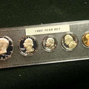 May include: A 1982 Year Set of U.S. coins in a clear plastic case. The set includes a half dollar, quarter, dime, nickel, and penny. The coins feature profiles of historical figures and are in excellent condition.