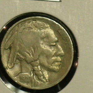 May include: A silver US Buffalo nickel coin with the year 1920 inscribed on the bottom. The coin features a profile of a Native American man with a feathered headdress.