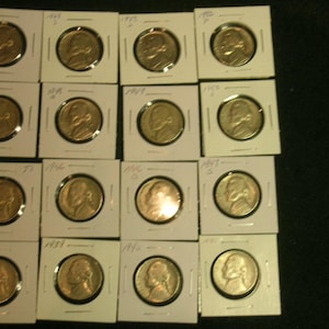 May include: A collection of vintage US nickel coins, each encased in a protective cardboard holder. The coins are a golden-bronze color, and the holders are white. The image shows a variety of dates, including 1953, 1949, and 1942.