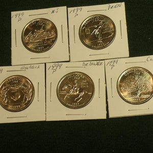 May include: Five 1999 P-mint mark United States state quarters in individual white cardboard holders. The coins feature designs for New Jersey, Pennsylvania, Georgia, Delaware, and Connecticut, with handwritten state names and dates on the holders.