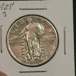 May include: A 1928 Standing Liberty quarter dollar coin encased in a white cardboard holder. The silver coin features the figure of Liberty holding a shield and olive branch, with the text LIBERTY, IN GOD WE TRUST, and the date 1928 visible.