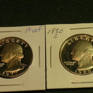 May include: Two silver-colored United States quarter coins in individual cardboard holders. The left coin is dated 1977 and the right coin is dated 1980. Both coins feature the profile of George Washington, the word LIBERTY, and IN GOD WE TRUST.