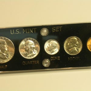 May include: A white ceramic mug with a black handle and the text "But First, Coffee" in a bold, black font. The mug is set against a blurred background, suggesting a cozy setting.