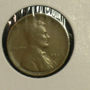 May include: A copper-colored coin with a profile of a man, the word "LIBERTY" and the year 1906 visible. The coin is encased in a clear plastic holder. A small, silver-colored pin is next to the coin.