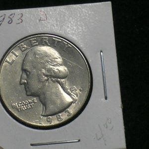 May include: A 1983 United States quarter coin, featuring George Washington's profile and the word "LIBERTY". The coin is held in a protective holder, with the year and a "D" mint mark visible. The words "IN GOD WE TRUST" are also visible.