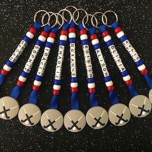 May include: A collection of personalized keychains with names spelled out in white letter beads. The keychains feature red, white, and blue beads, blue ribbons, and gray hockey-themed charms with crossed hockey sticks. Each keychain has a silver keyring.