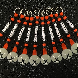 May include: A collection of personalized keychains with names spelled out in white letter beads. Each keychain features a gray disc with a hockey stick and ball design, connected by orange ribbon and black and orange beads.
