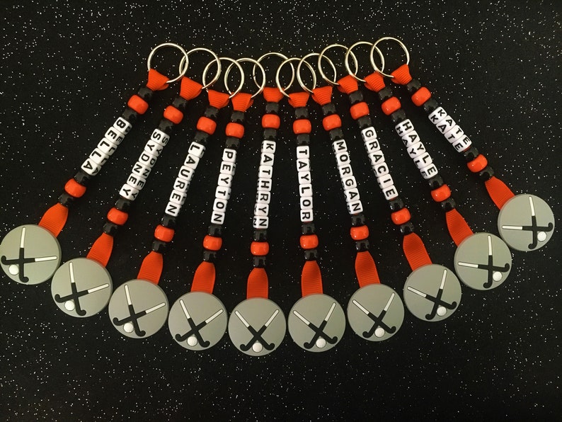 May include: A collection of personalized keychains with names spelled out in white letter beads. Each keychain features a gray disc with a hockey stick and ball design, connected by orange ribbon and black and orange beads.