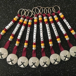 May include: A collection of personalized hockey keychains. Each keychain features a round gray charm with crossed hockey sticks, a maroon ribbon, and a beaded strand with a name spelled out in white letter beads. The keychains are in a variety of colors.