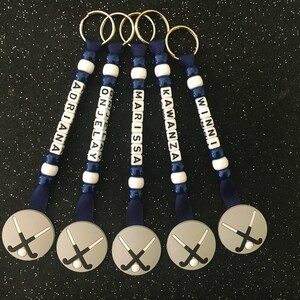 May include: Five personalized keychains with names spelled out in white letter beads. The keychains have a navy blue and white bead design, navy blue ribbon, and a silver charm with crossed hockey sticks. Names include: Adriana, Onjelay, Marissa, Kawanza, and Winni.