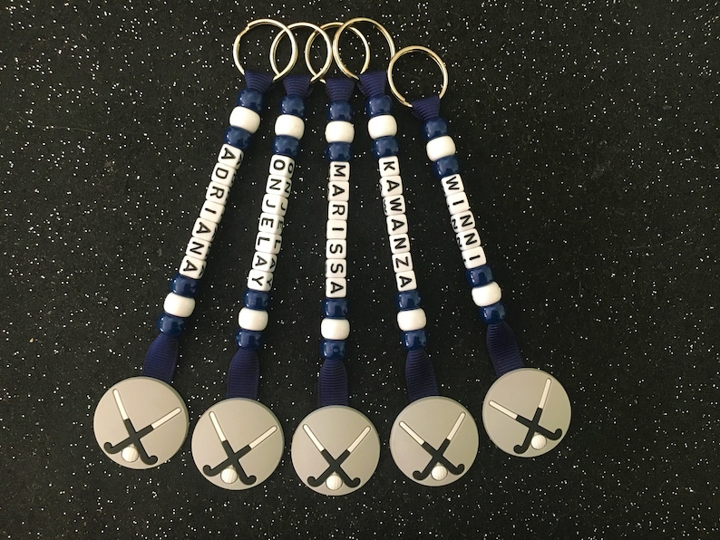 May include: Five personalized keychains with names spelled out in white letter beads. The keychains have a navy blue and white bead design, navy blue ribbon, and a silver charm with crossed hockey sticks. Names include: Adriana, Onjelay, Marissa, Kawanza, and Winni.