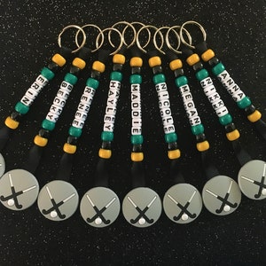 May include: A collection of personalized keychains with names spelled out in white letter beads. Each keychain features a hockey stick charm and is accented with green, black, and gold beads. The names include Erin, Becky, Renee, Hayley, Maddie, Nicole, Megan, Anna, and Nikki.