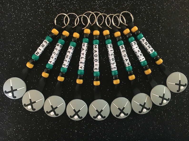 May include: A collection of personalized keychains with names spelled out in white letter beads. Each keychain features a hockey stick charm and is accented with green, black, and gold beads. The names include Erin, Becky, Renee, Hayley, Maddie, Nicole, Megan, Anna, and Nikki.