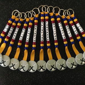 May include: A collection of personalized keychains with names spelled out on beaded strands. Each keychain features a gray circular charm with crossed hockey sticks and a yellow ribbon.