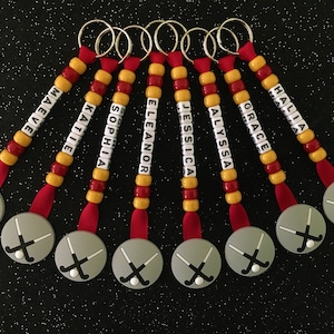 May include: A collection of personalized hockey keychains. Each keychain features a gray disc with crossed hockey sticks, a red ribbon, and a beaded strand with a name spelled out in white letter beads. The beads are red and yellow.