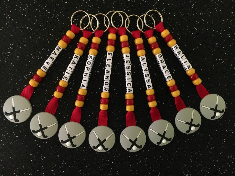 May include: A collection of personalized hockey keychains. Each keychain features a gray disc with crossed hockey sticks, a red ribbon, and a beaded strand with a name spelled out in white letter beads. The beads are red and yellow.