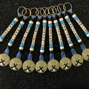 May include: A collection of personalized keychains with names spelled out in white letter beads. Each keychain has a blue ribbon, gold ring, and a circular charm with crossed hockey sticks. The names include: Shayne, Roarie, Madison, Abigail, McKenzie, Emerson, Ryleigh, Addison, Dallilah, and Austin.