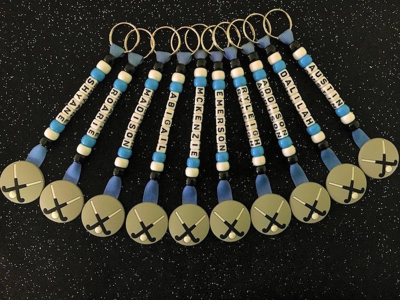 May include: A collection of personalized keychains with names spelled out in white letter beads. Each keychain has a blue ribbon, gold ring, and a circular charm with crossed hockey sticks. The names include: Shayne, Roarie, Madison, Abigail, McKenzie, Emerson, Ryleigh, Addison, Dallilah, and Austin.