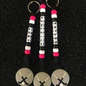 May include: Three personalized keychains with black, white, and pink beads spelling out names. Each keychain has a gray hockey puck charm with crossed hockey sticks and a ball. The keychains are on a black glitter background.