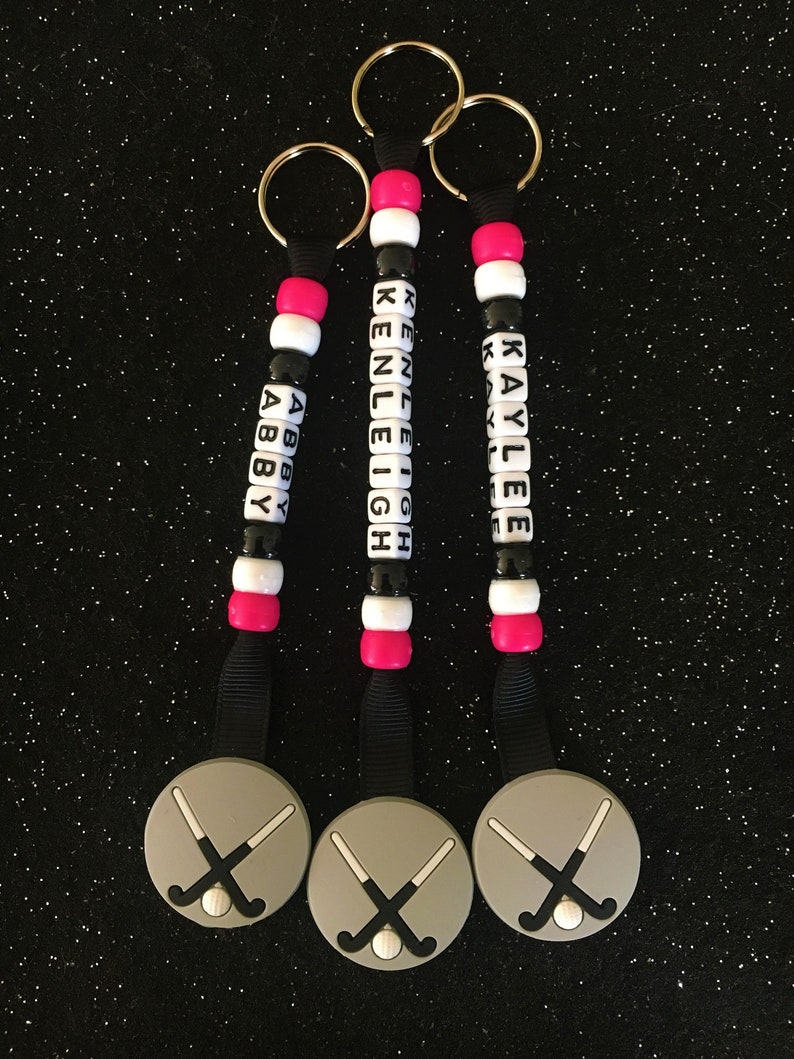 May include: Three personalized keychains with black, white, and pink beads spelling out names. Each keychain has a gray hockey puck charm with crossed hockey sticks and a ball. The keychains are on a black glitter background.