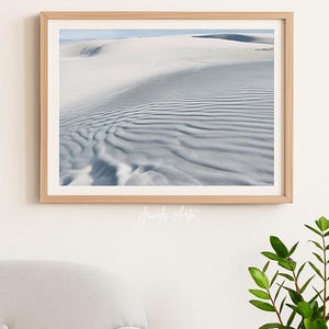 May include: A framed print featuring a landscape of white sand dunes with rippled textures under a blue sky. The artwork is in a light wood frame and hangs on a white wall. The text "Jewels Arte" is in the bottom center of the print.