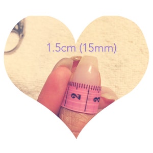 May include: A close-up of a finger with a pink measuring tape wrapped around it. The tape reads "2" and "3". The text "1.5cm (15mm)" is written above the finger.