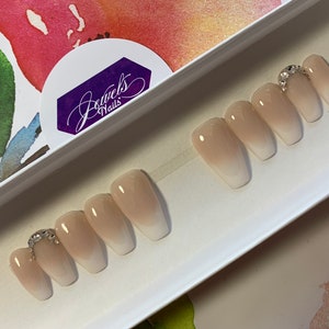 May include: Set of five nude-colored coffin-shaped press-on nails with a gradient ombre effect. The nails have a clear base with a light pink color at the tips. The nails are decorated with a single row of rhinestones on the top of the nail. The nails are displayed in a white box with the text "Jewels Nails" on the lid.