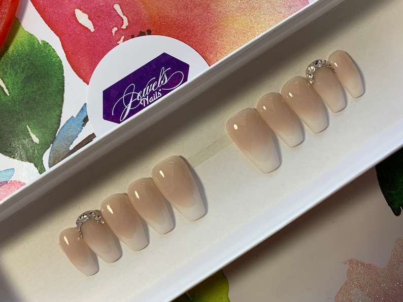 May include: Set of five nude-colored coffin-shaped press-on nails with a gradient ombre effect. The nails have a clear base with a light pink color at the tips. The nails are decorated with a single row of rhinestones on the top of the nail. The nails are displayed in a white box with the text "Jewels Nails" on the lid.