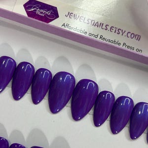 Puede incluir: Un conjunto de diez u&ntilde;as postizas de color morado con un acabado brillante. Las u&ntilde;as est&aacute;n dispuestas en una fila sobre una superficie blanca. JEWELSNAILS.ETSY.COM est&aacute; impreso en una caja blanca en el fondo. Presiona sobre asequible y reutilizable.